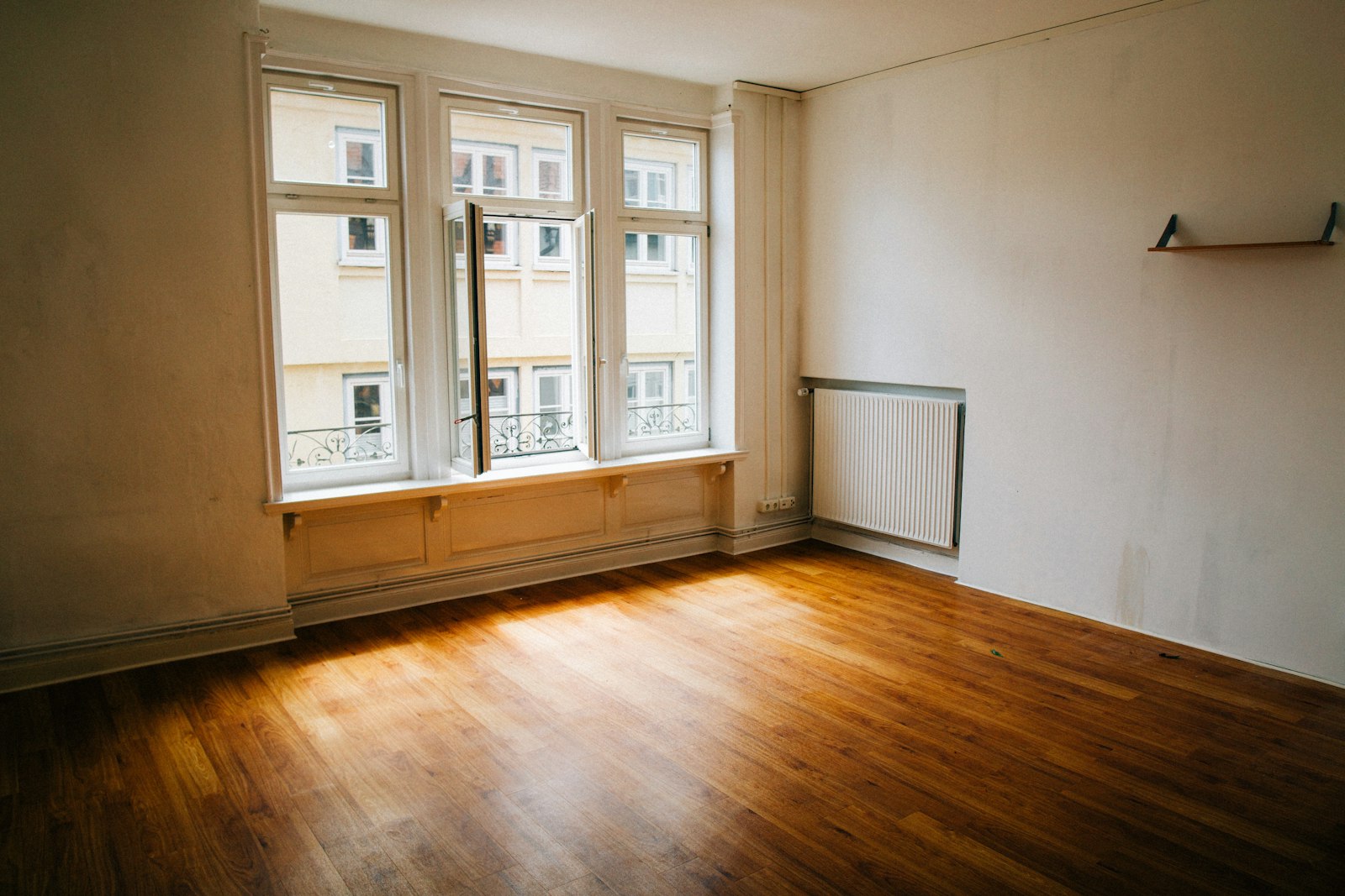 Empty apartment room with wood floor, tall windows, and a radiator